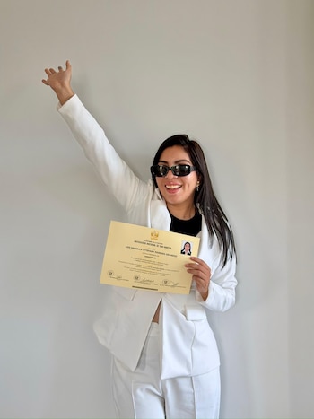 Mujer con traje blanco y gafas de sol oscuras sostiene un diploma profesional dorado y levanta el brazo derecho, frente a una pared lisa