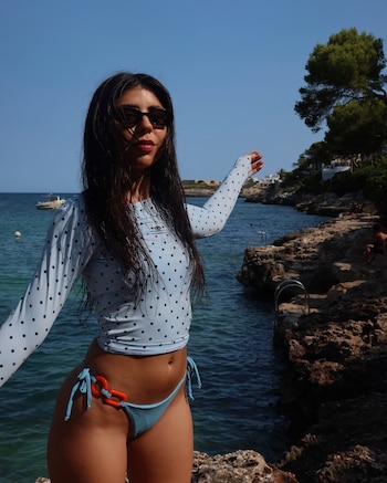 Mujer con cabello oscuro y gafas de sol, vistiendo traje de baño celeste de lunares, posa junto al mar en una cala rocosa con un barco