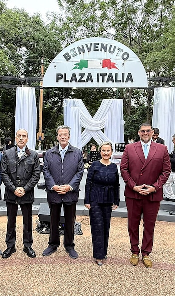 Cuatro personas, tres hombres y una mujer, de pie al frente de un escenario blanco con un letrero "Benvenuto Plaza Italia" en un evento al aire libre