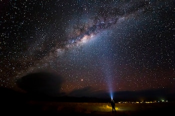 Cielo nocturno con estrellas y la Vía Láctea sobre un terreno oscuro donde una persona apunta una linterna hacia arriba