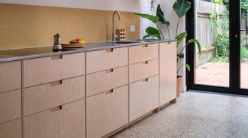 Cocina moderna con gabinetes de madera clara, encimera de acero inoxidable, azulejos amarillos, fregadero y cafetera. Puerta de cristal a jardín.