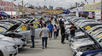 Mercado de autos usados al aire libre. Filas de vehículos con capós abiertos, personas inspeccionando. Banderolas de venta y edificios urbanos al fondo.