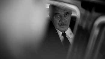 Retrato en blanco y negro de un hombre mayor con bigote, vistiendo traje y corbata, sentado en un vehículo y mirando fijamente. Elementos del interior del vehículo se ven borrosos en primer plano