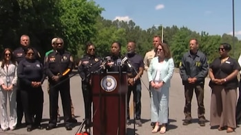 Una oficial de policía en uniforme negro habla en un podio con micrófonos, flanqueada por varios oficiales y personas vestidas de civil bajo un cielo azul claro