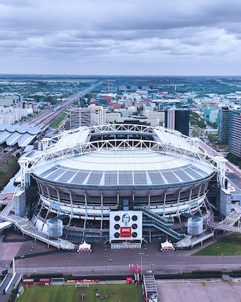 Vista aérea del Johan Cruijff Arena en Ámsterdam con techo retráctil y paneles solares; banner de Johan Cruijff, campo de fútbol y ciudad al fondo