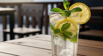 Primer plano de un vaso de agua con hielo, rodajas de limón y hojas de hierbabuena sobre una mesa de madera. El vaso está cubierto de condensación.