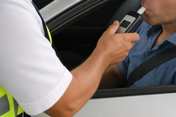 Un agente con chaleco reflectante sostiene un etilómetro digital en la boca de un hombre sentado al volante de un automóvil.