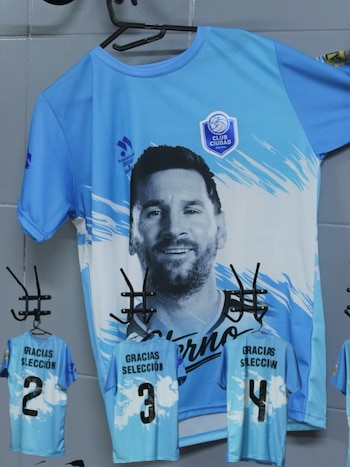 Una camiseta de fútbol celeste con la cara de Lionel Messi está colgada, junto a tres camisetas infantiles celestes con los números dos, tres y cuatro