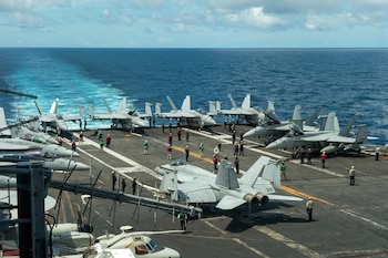 Varios aviones de combate grises y tripulantes con chalecos de colores se encuentran sobre la cubierta de un portaaviones en pleno mar