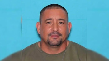 Primer plano frontal de un hombre adulto con barba y bigote, cabeza rapada, vistiendo una camiseta verde oliva, sobre un fondo azul claro