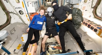Tres personas flotan en un módulo espacial; una lleva una máscara de extraterrestre y otra viste un pasamontañas negro