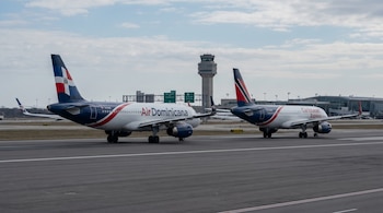 Dos aviones comerciales, uno con la bandera de República Dominicana y otro con la de Trinidad y Tobago, en paralelo sobre una pista de aeropuerto, con una torre de control al fondo.
