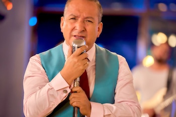 Hombre maduro con camisa rosa, chaleco celeste y corbata roja sostiene un micrófono clásico metálico con ambas manos ante el rostro