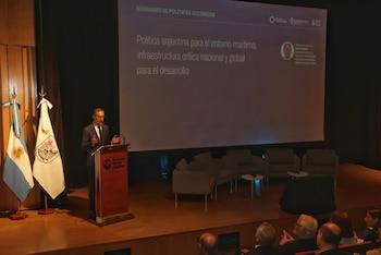 Un hombre habla desde un atril junto a dos banderas frente a una gran pantalla de proyección en un auditorio