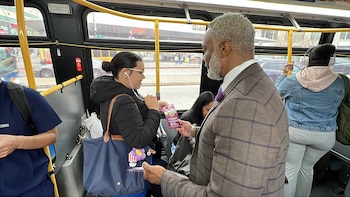 Um homem barbudo de terno entregando um folheto roxo para uma mulher de óculos e fones de ouvido em um ônibus. Outro passageiro na parte de trás
