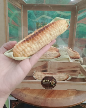 Una mano sostiene un pan horneado con forma de mazorca. Otros panes similares se encuentran en una caja expositora de madera con un logo de Bachi