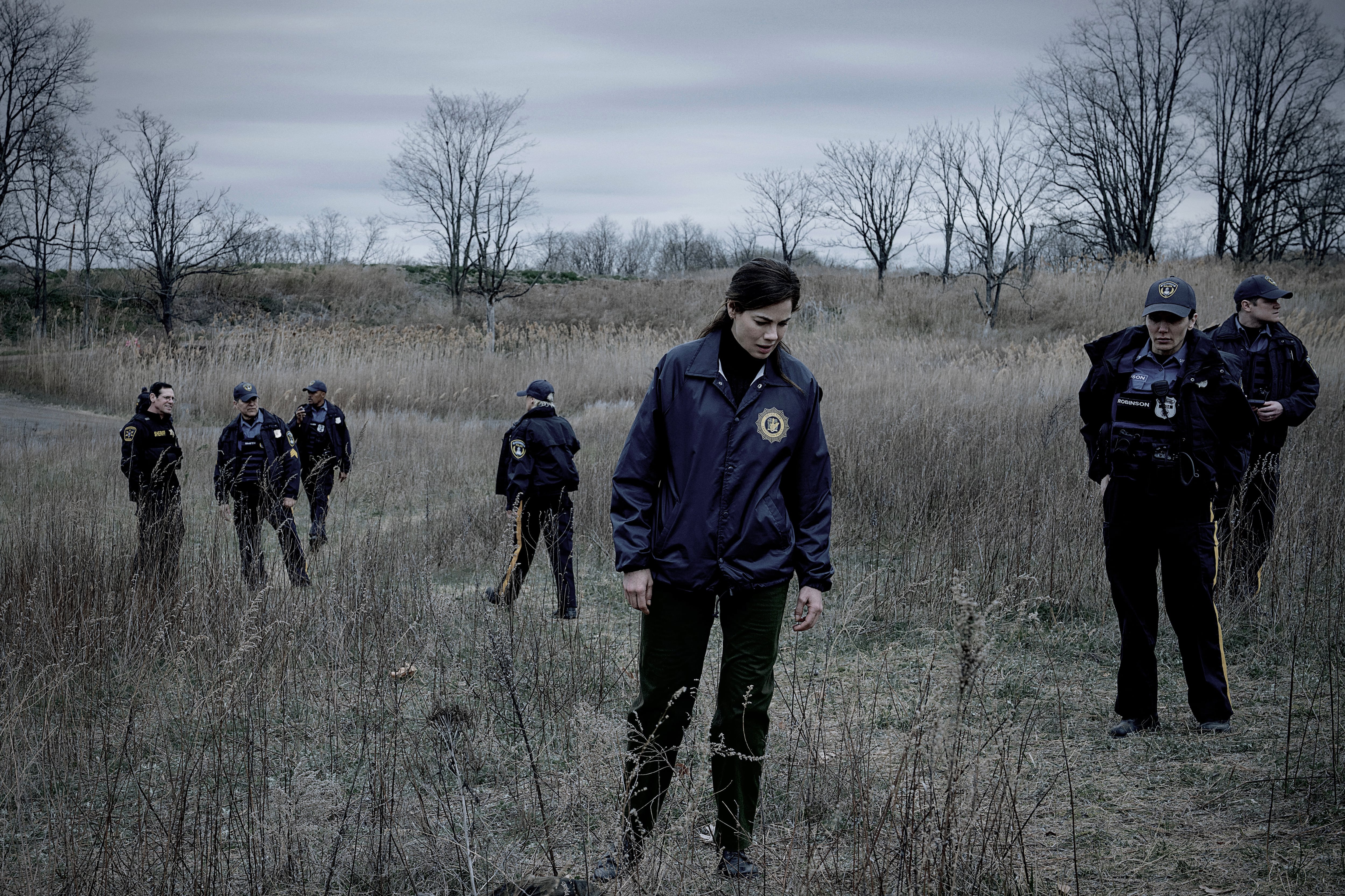 La actriz Michelle Monaghan, interpretando a una oficial de policía, avanza por un campo de hierba seca junto a otros agentes policiales bajo un cielo nublado. (Netflix)