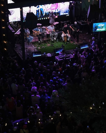 Músicos con guitarras tocan en un escenario con pantallas y luces frente a un grupo numeroso de personas en un recinto al aire libre