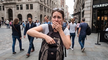 Una mujer con mochila, gafas de sol en la cabeza y camiseta de rayas, tiene una mano en la boca y la otra dentro de la mochila en una calle con edificios y gente.