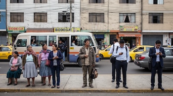 Personas con diferentes vestimentas esperan en una acera junto a una calle transitada por taxis amarillos, un minibús y edificios comerciales.