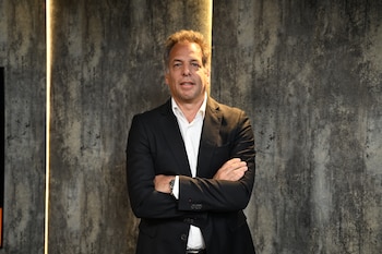 Un hombre de cabello castaño claro viste un saco negro y camisa blanca con los brazos cruzados ante un fondo gris texturizado