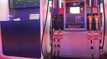 Surtidor de combustible con cuatro mangueras negras y amarillas, pantalla digital con números, y rótulos de nafta y gasoil. Un mostrador con laptop y dispositivo blanco.