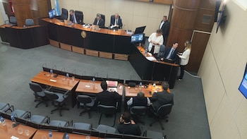 Vista aérea de una sala de audiencias moderna con varias personas sentadas en un estrado curvo oscuro y mesas claras. Algunas personas están de pie. Bandera argentina visible