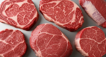 Primer plano de seis cortes de carne vacuna cruda en tonos rojos y blancos, sobre una superficie de acero inoxidable gris claro.