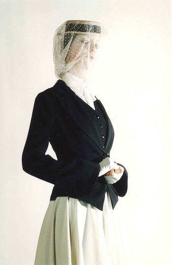 Una modelo o maniquí de la cintura para arriba, lleva una chaqueta negra, blusa blanca, falda, un sombrero con velo de lunares y guantes blancos