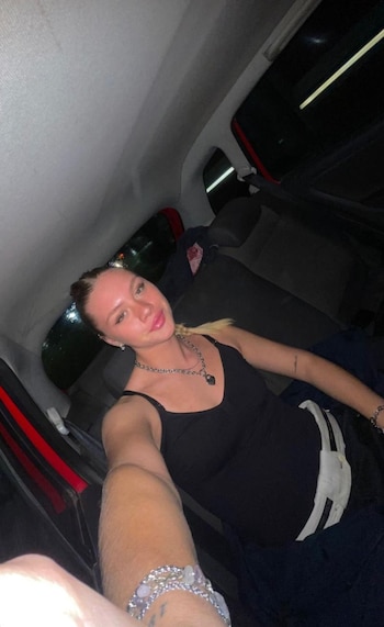 Mujer joven con cabello rubio atado y una coleta, viste un top negro, collar y pulsera, sonríe en el asiento trasero de un automóvil oscuro