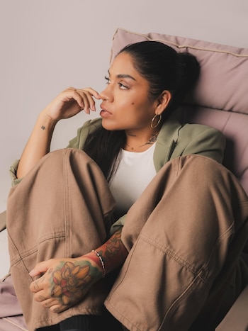 Massiel Málaga sentada en un sillón rosa, con blazer verde oliva y pantalones marrones, mira pensativa hacia la derecha. Se ven sus tatuajes en los brazos