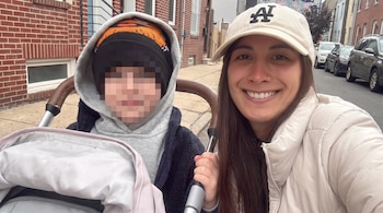 Mujer con gorra y campera clara junto a un niño con gorro y capucha en cochecito. La cara del niño está pixelada. Hay edificios de ladrillo y coches estacionados detrás.