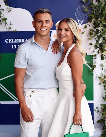 Un hombre y una mujer jóvenes, ambos sonrientes, posan de frente a la cámara. El hombre viste polo azul claro y pantalón blanco; la mujer, vestido blanco con bolso verde