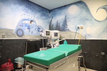 Interior de un quirófano con paredes pintadas con un mural de cielo nocturno, un coche y elementos flotantes. Una camilla verde y equipo médico están en el centro