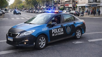 Un automóvil patrullero de la Policía de la Ciudad de Buenos Aires con balizas encendidas en una calle urbana con edificios, árboles y personas.