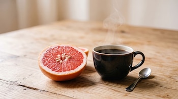Taza de café humeante, medio pomelo rosado, cuchara plateada, mesa de madera clara.