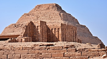 Una pirámide escalonada de color ocre con estructuras de piedra antiguas en primer plano, bajo un cielo azul sin nubes.