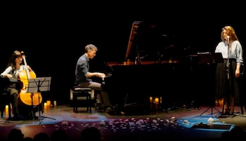 Tres músicos actúan en un escenario oscuro: una mujer toca el chelo, un hombre el piano y otra mujer canta. Velas y objetos blancos adornan el suelo