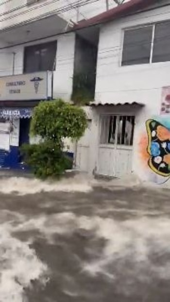 Calle con agua de inundación, dos edificios, un consultorio médico con rótulo, una puerta blanca, un árbol y un grafiti de mariposa en una pared