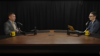 Dos hombres sentados en una mesa de madera con micrófonos, cada uno con una taza amarilla, frente a un telón de fondo negro durante una grabación