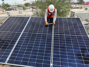 Un trabajador con casco y chaleco naranja instala paneles solares azules en un tejado. Se observan otros tejados y unidades de aire acondicionado al fondo