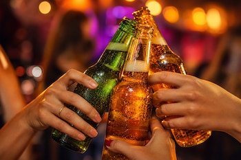 Manos sostienen tres botellas de cerveza de vidrio verde y ámbar en un brindis sobre un fondo de luces desenfocadas.