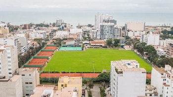 Vista aérea de una ciudad costera con grandes campos deportivos de césped y canchas de tenis de arcilla, rodeados de edificios y el mar al fondo