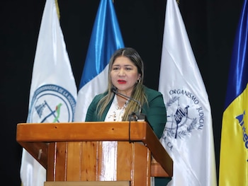 Una mujer con chaqueta verde habla frente a un micrófono en un podio de madera, con varias banderas en el fondo