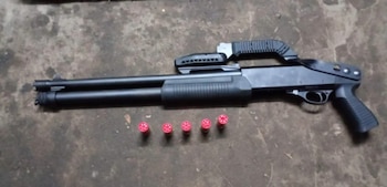 Una escopeta negra de estilo táctico con una empuñadura de pistola y cuatro cartuchos rojos de escopeta alineados sobre una superficie oscura