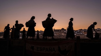 Siluetas de varias mujeres, algunas con sacos, sobre una estructura de madera. Al fondo, un pueblo o ciudad y el cielo al amanecer. Una bandera española cuelga al frente.