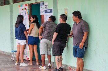 Un grupo de cinco personas de pie junto a una pared verde claro y dos puertas, con carteles electorales visibles. Algunas personas conversan, otras esperan