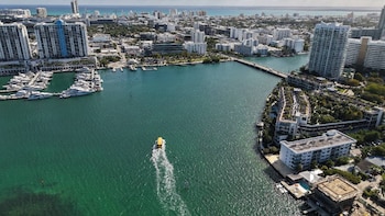Vista aérea de una ciudad costera con edificios altos, un puerto deportivo lleno de barcos, un puente y un bote amarillo navegando por el agua