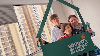 Una mujer y dos jóvenes sostienen un marco verde con forma de casa con la inscripción Bogotá mi ciudad mi casa