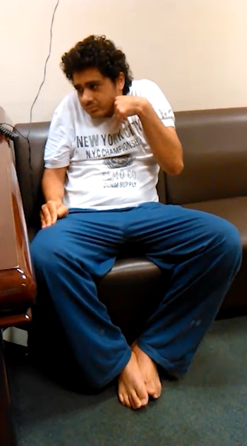 Un hombre descalzo con camiseta blanca y pantalón azul permanece sentado en un sofá de cuero marrón junto a un mueble